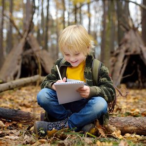 Little boy scout is orienteering in forest. Child is sitting on fallen tree and writing in the notepad. Behind the child is teepee hut.