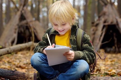 Little boy scout is orienteering in forest. Child is sitting on fallen tree and writing in the notepad. Behind the child is teepee hut.