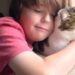 A young boy with light brown hair, wearing a pink shirt, is seen in a close-up hugging a brown, white, and black tabby cat. The boy has his eyes closed and a soft smile, resting his head against the cat. The cat, with prominent green eyes and long white whiskers, looks contentedly out of a window to the right of the frame, bathed in natural light.