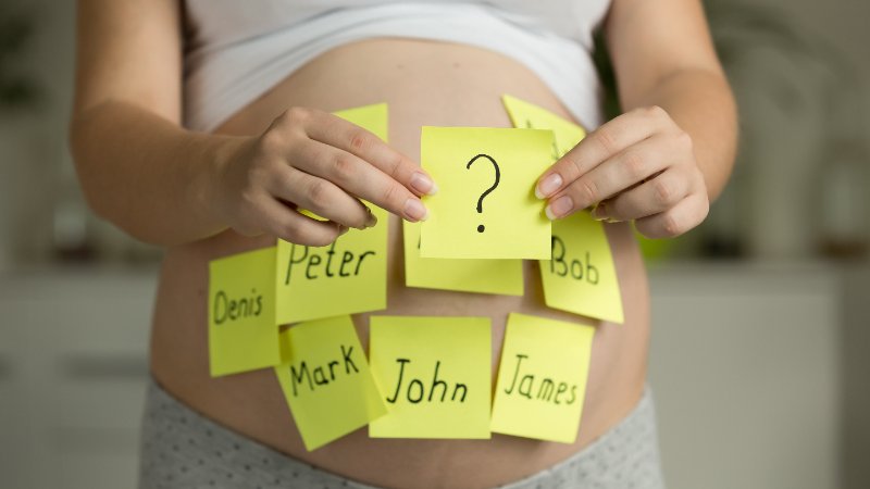 A close-up view of a pregnant woman's bare abdomen adorned with several yellow sticky notes, each bearing a handwritten male name such as 'Peter', 'Denis', 'Mark', 'John', 'James', and 'Bob'. Her hands gently hold a central sticky note prominently displaying a black question mark, symbolizing the thoughtful process of selecting a baby name. She wears a white top and gray patterned bottoms, with a softly blurred background.