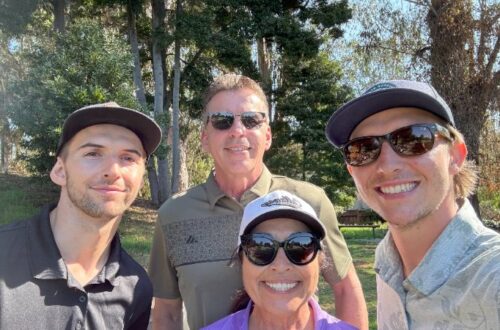 A group of four smiling people are posing outdoors on a sunny day. From left to right, a young man with a short beard wearing a black cap and shirt, an older man with short brown hair and sunglasses wearing an olive green polo shirt, a woman with dark hair and sunglasses wearing a white cap and purple top, and a young man with a slight beard wearing a blue cap and sunglasses with a light patterned short-sleeved shirt. They are standing in a grassy area with tall green trees and leafy foliage in the background.