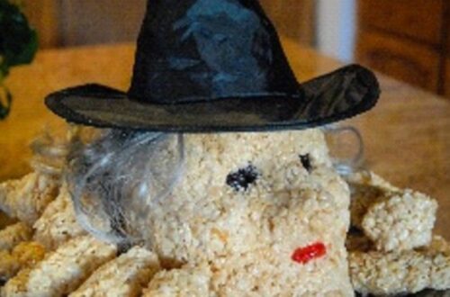 A close-up shows a homemade Halloween treat shaped like a witch's head, made from light brown puffed cereal. The witch wears a black pointed hat and has wispy grey 'hair' around its base. Its face features two small black dots for eyes and a small red candy for lips. Additional arms or body parts made of the same cereal extend from the witch's head.