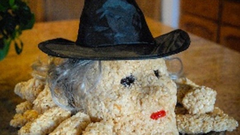 A close-up shows a homemade Halloween treat shaped like a witch's head, made from light brown puffed cereal. The witch wears a black pointed hat and has wispy grey 'hair' around its base. Its face features two small black dots for eyes and a small red candy for lips. Additional arms or body parts made of the same cereal extend from the witch's head.