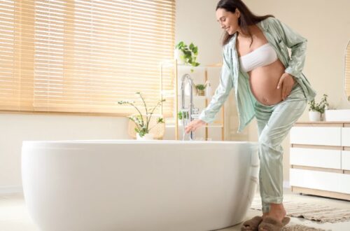 A smiling pregnant woman, wearing light green satin pajamas and a white bandeau top, leans over a modern white freestanding bathtub. She is gently touching the running water from a chrome faucet with her right hand, while her left hand is cradling her baby bump. The bright bathroom features light wood blinds on a window, potted green plants, a white vanity, and a round mirror. She is wearing fuzzy brown slippers on a striped bath mat.