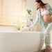 A smiling pregnant woman, wearing light green satin pajamas and a white bandeau top, leans over a modern white freestanding bathtub. She is gently touching the running water from a chrome faucet with her right hand, while her left hand is cradling her baby bump. The bright bathroom features light wood blinds on a window, potted green plants, a white vanity, and a round mirror. She is wearing fuzzy brown slippers on a striped bath mat.