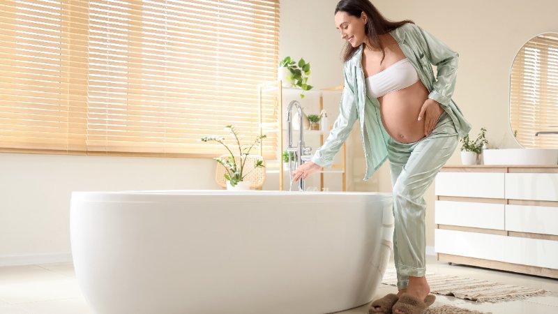 A smiling pregnant woman, wearing light green satin pajamas and a white bandeau top, leans over a modern white freestanding bathtub. She is gently touching the running water from a chrome faucet with her right hand, while her left hand is cradling her baby bump. The bright bathroom features light wood blinds on a window, potted green plants, a white vanity, and a round mirror. She is wearing fuzzy brown slippers on a striped bath mat.
