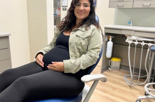 A smiling pregnant woman with dark, curly hair and glasses sits comfortably in a blue dental chair. She is wearing a black tank top, black leggings, and a light green corduroy jacket, with her hands gently resting on her visibly pregnant belly. The background shows a clean dental office setting with grey cabinets, dental equipment, and a bottle of hand sanitizer on a counter.