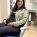 A smiling pregnant woman with dark, curly hair and glasses sits comfortably in a blue dental chair. She is wearing a black tank top, black leggings, and a light green corduroy jacket, with her hands gently resting on her visibly pregnant belly. The background shows a clean dental office setting with grey cabinets, dental equipment, and a bottle of hand sanitizer on a counter.