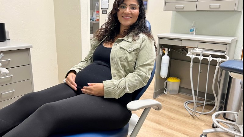 A smiling pregnant woman with dark, curly hair and glasses sits comfortably in a blue dental chair. She is wearing a black tank top, black leggings, and a light green corduroy jacket, with her hands gently resting on her visibly pregnant belly. The background shows a clean dental office setting with grey cabinets, dental equipment, and a bottle of hand sanitizer on a counter.