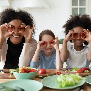 In kitchen couple and multi racial daughters prepare vegetable salad having fun make funny faces cover eyes with red paprika circles looking like eyewear, binoculars shape, cookery, family joy concept